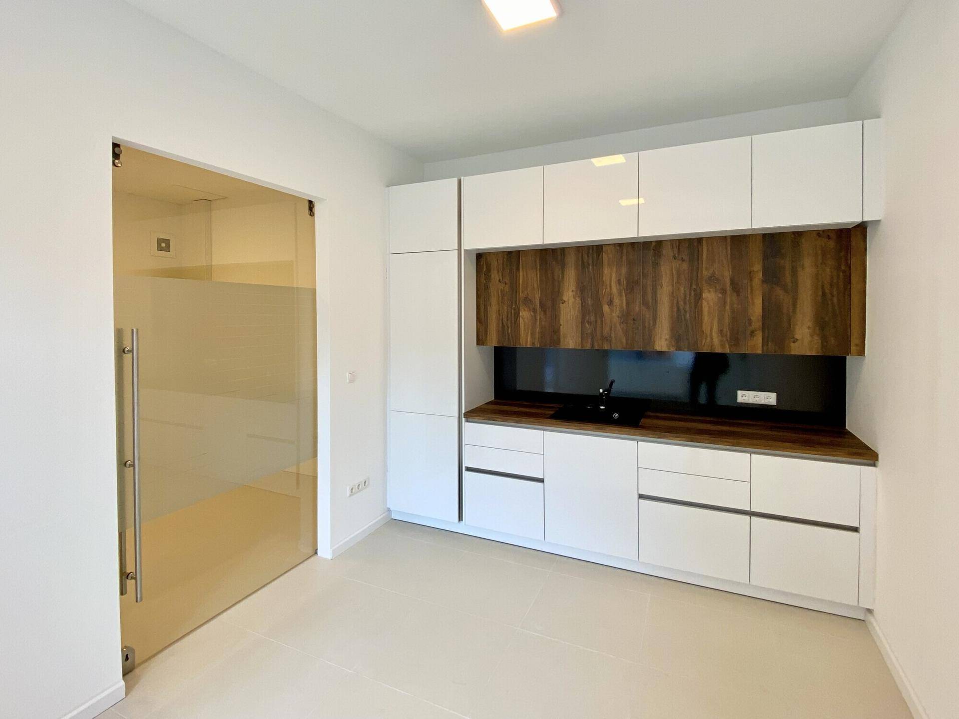 Moderne Küchenzeile mit weißen Hochglanzfronten und Holzdetails, durch eine Glastür abgetrennt.