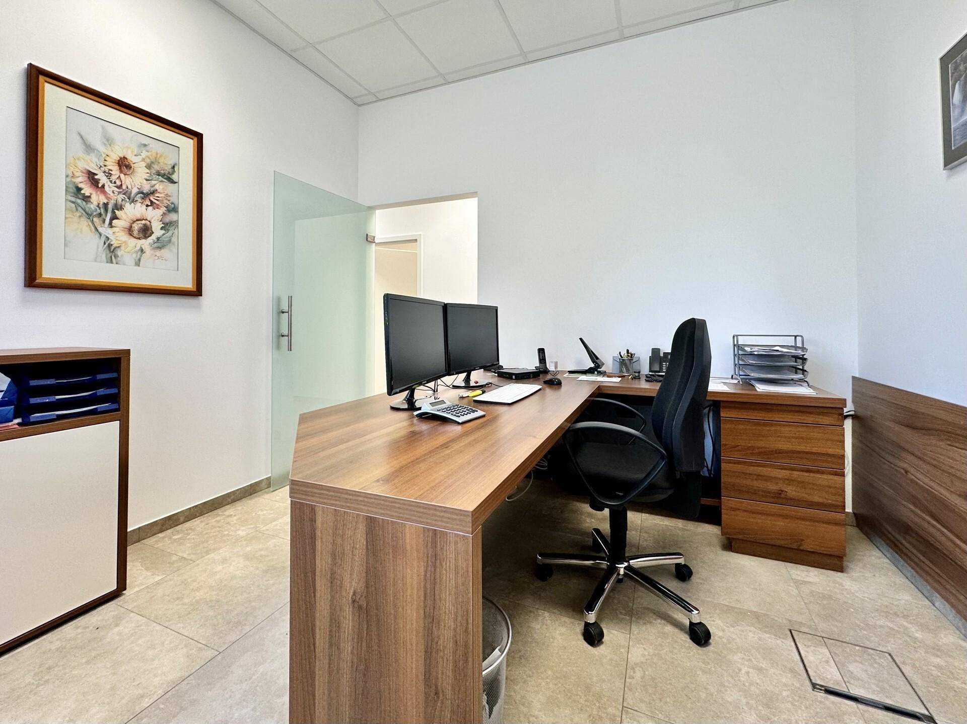 Büro mit einem L-förmigen Holzschreibtisch, Monitoren und einer Glastür, die zu einem weiteren Raum führt.