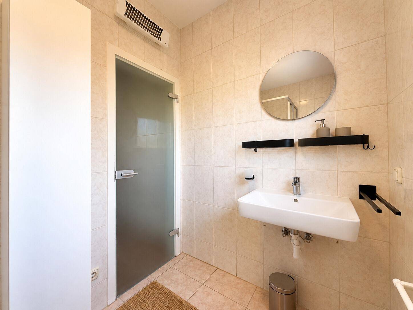 Badezimmer mit modernem Waschbecken, rundem Spiegel und einer stilvollen Glastür.