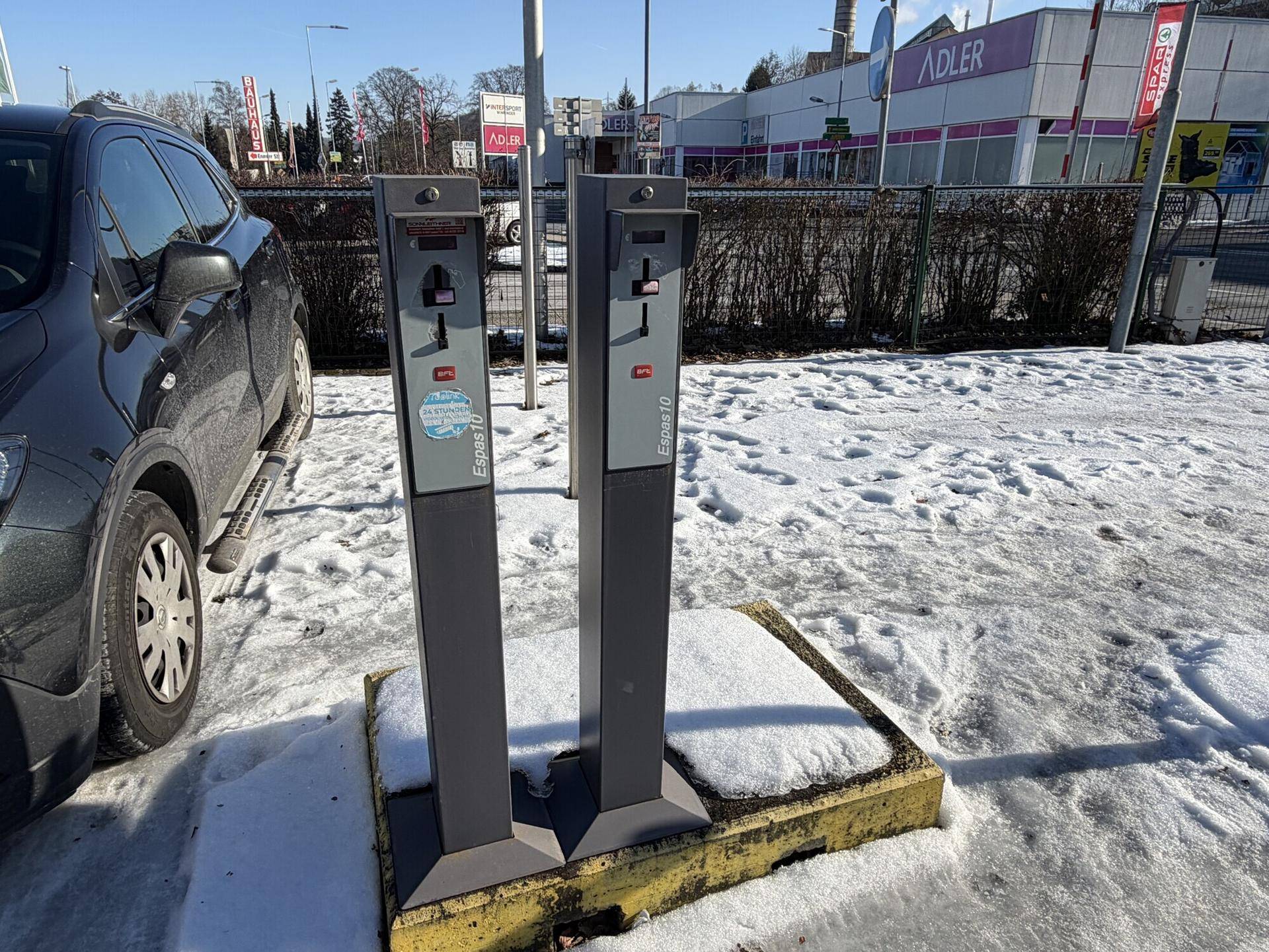 Zwei Parkautomaten stehen auf einem verschneiten Parkplatz neben einem geparkten Auto.