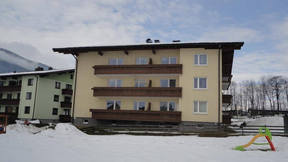 Mehrfamilienhaus mit Balkonen und einem kleinen Spielgerät im verschneiten Gartenbereich mit Bergblick.