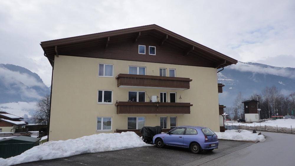 Ansicht des Mehrfamilienhauses mit Balkonen, einem geparkten Auto und verschneiter Umgebung vor Bergkulisse.