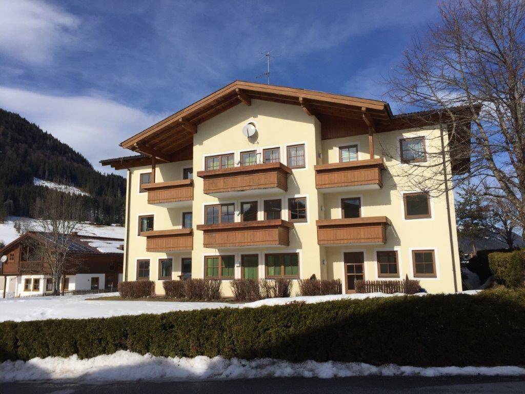 Alpines Wohngebäude mit gelber Fassade, Holzbalkonen und schneebedeckter Umgebung vor Bergkulisse.