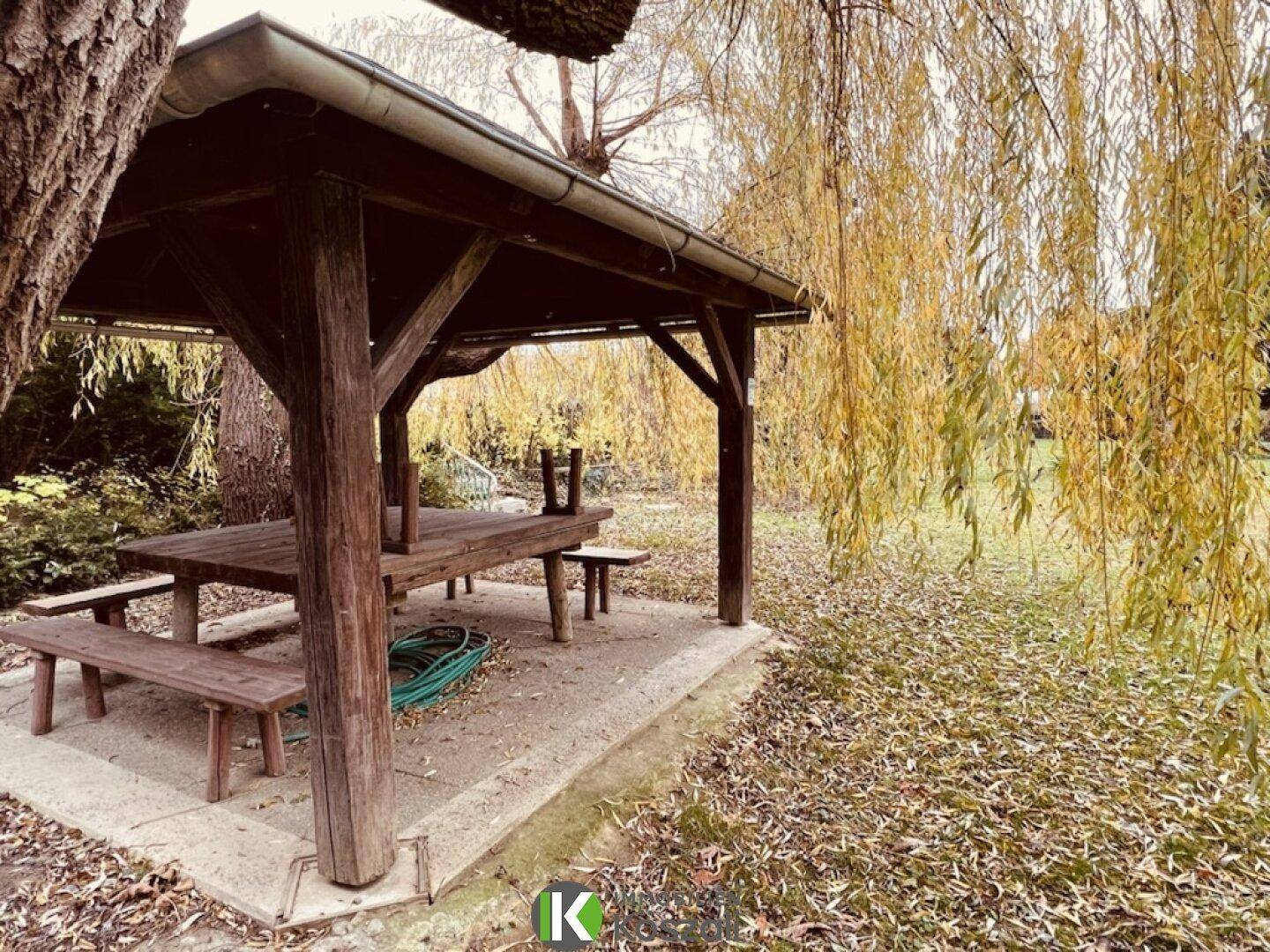 Gemütlicher Holzpavillon mit Picknicktisch im Garten, umgeben von Bäumen und Herbstlaub.