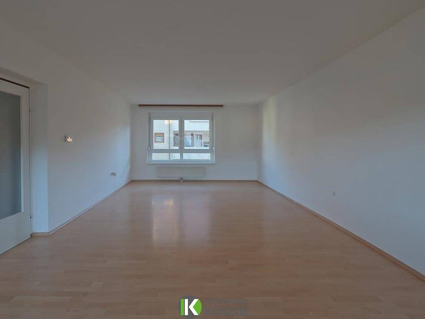 Geräumiges, leeres Wohnzimmer mit hellem Holzboden und großem Fenster, ideal für geselliges Beisammensein.