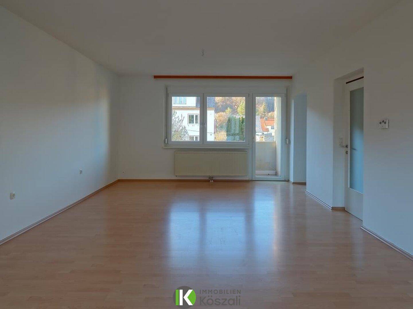 Geräumiges, leeres Wohnzimmer mit hellem Holzboden, großem Fenster und Zugang zum Balkon.