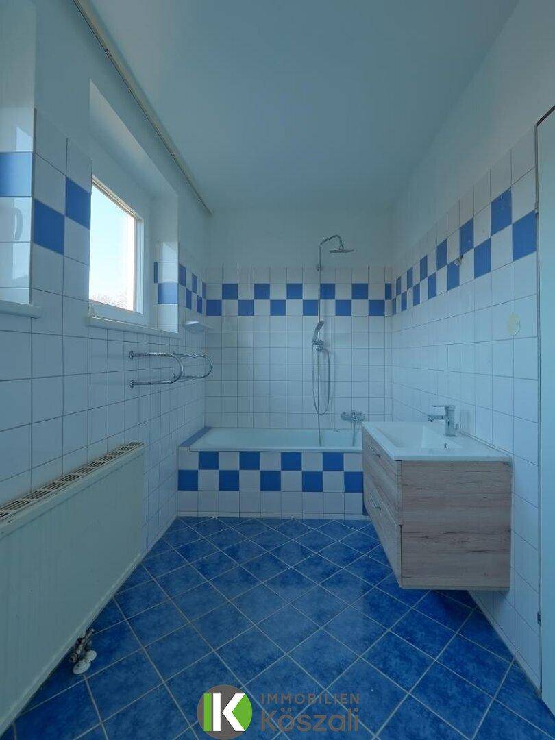 Badezimmer mit blauen und weißen Fliesen, Badewanne mit Duschfunktion und Fenster für Tageslicht.