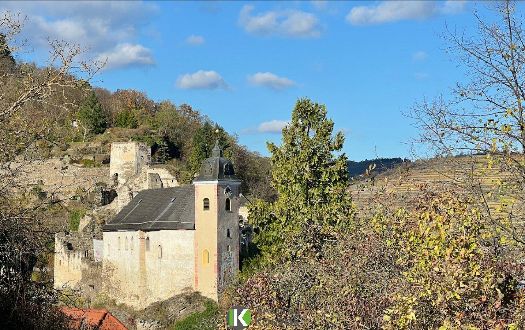 Malerischer Blick auf eine historische Kirche und Ruinen inmitten grüner Hügel unter blauem Himmel.