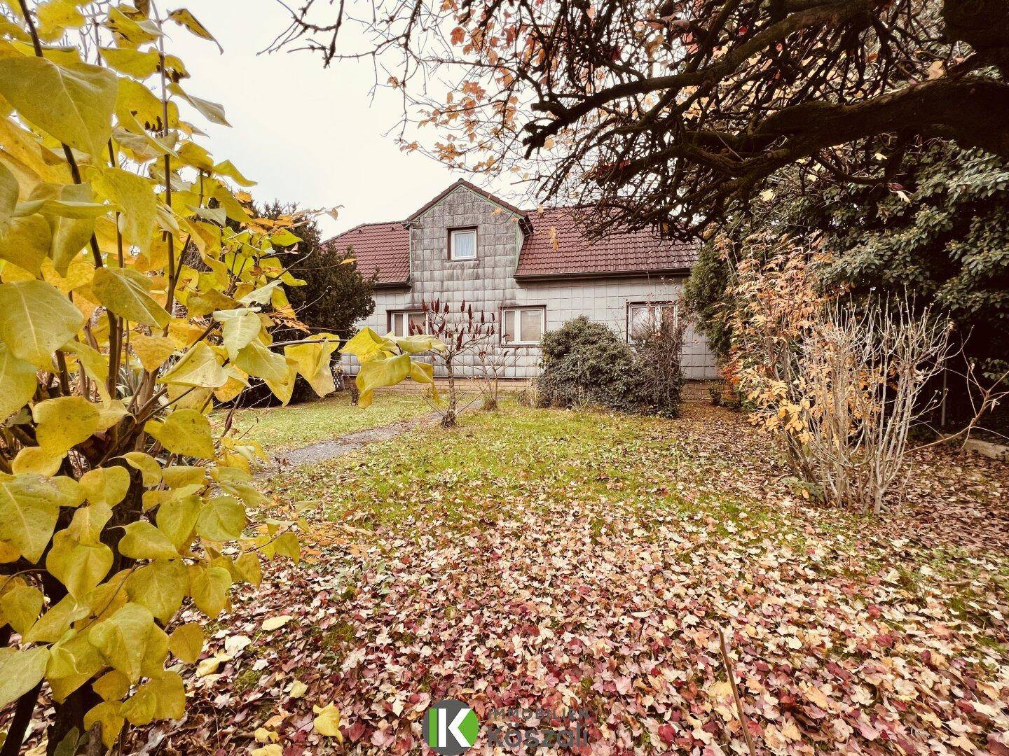 Vorderansicht eines Hauses mit Garten und herbstlicher Bepflanzung, umgeben von Bäumen.