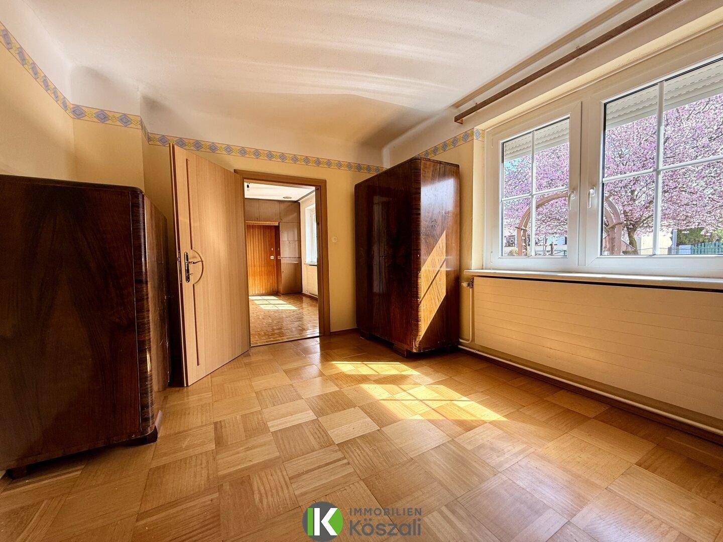 Helles Schlafzimmer mit Parkettboden, Holzschränken und Blick auf einen blühenden Baum.