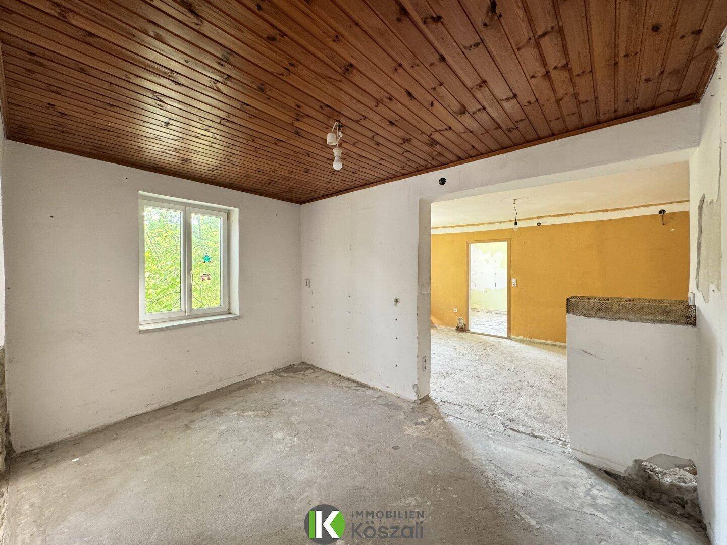 Renovierungsbedürftiger Raum mit Holzbalkendecke und Betonboden, offen zu einem weiteren Bereich.