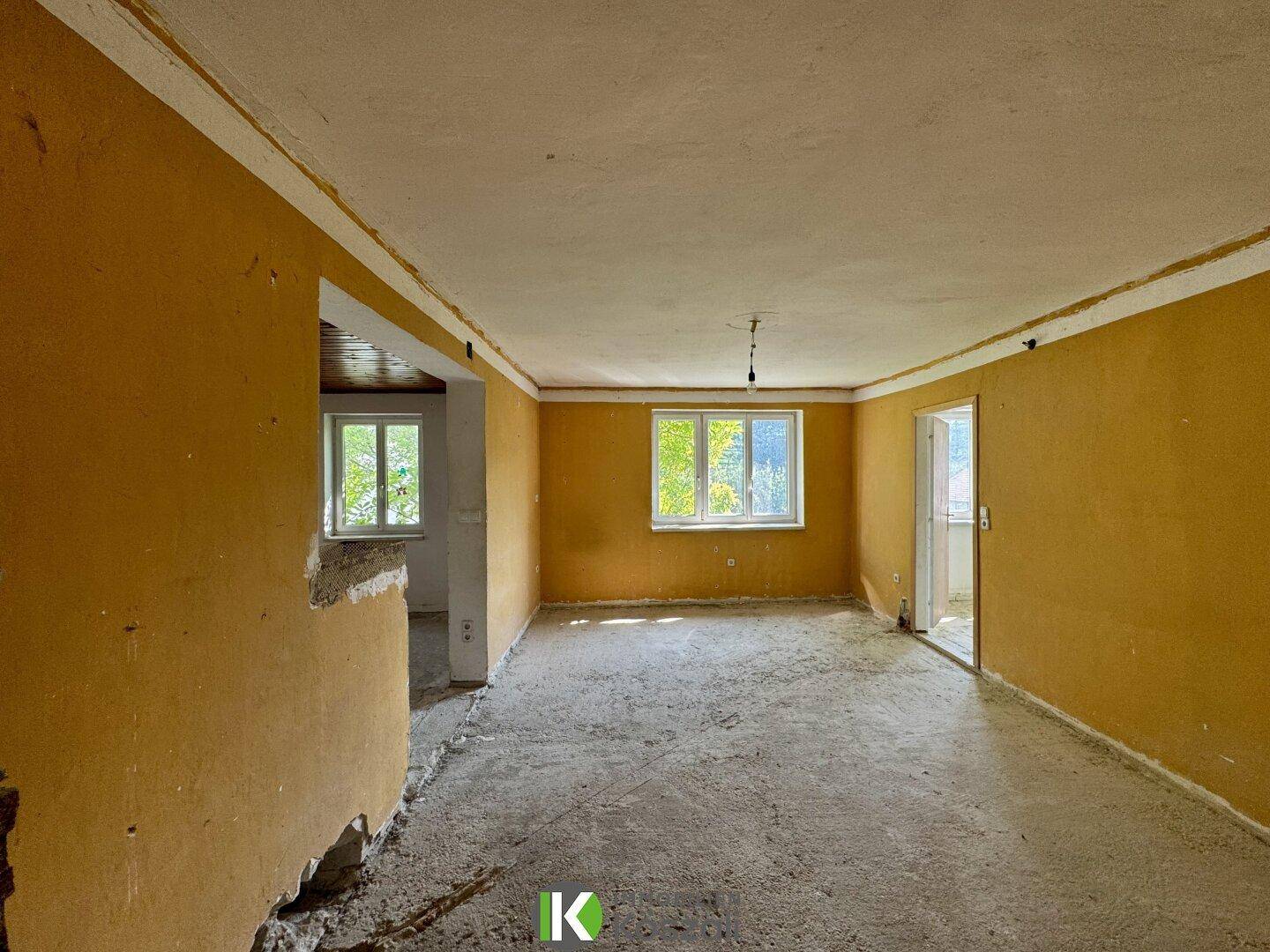 Renovierungsbedürftiger Raum mit gelben Wänden und Betonboden, offen zu einem weiteren Bereich.