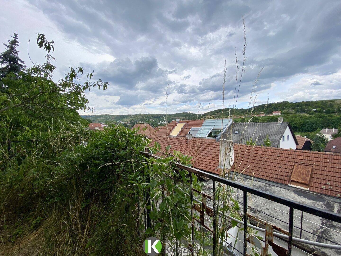 Blick von einem Balkon über die Dächer der Umgebung und die grüne Landschaft unter bewölktem Himmel.