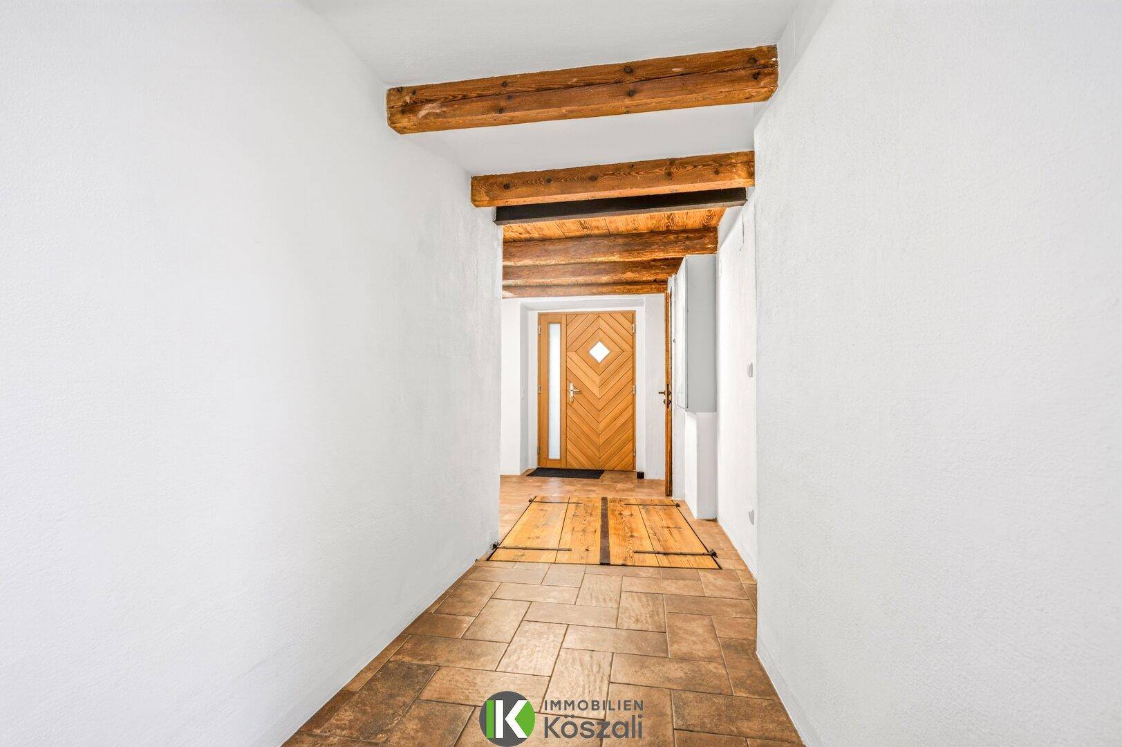 Eingangsbereich mit Holztür, Terrakottafliesen und sichtbaren Holzbalken, der zu weiteren Räumen führt.