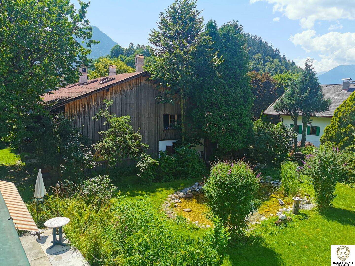 Weitläufiger Garten mit Teich und Blick auf die umliegende Berglandschaft unter blauem Himmel.