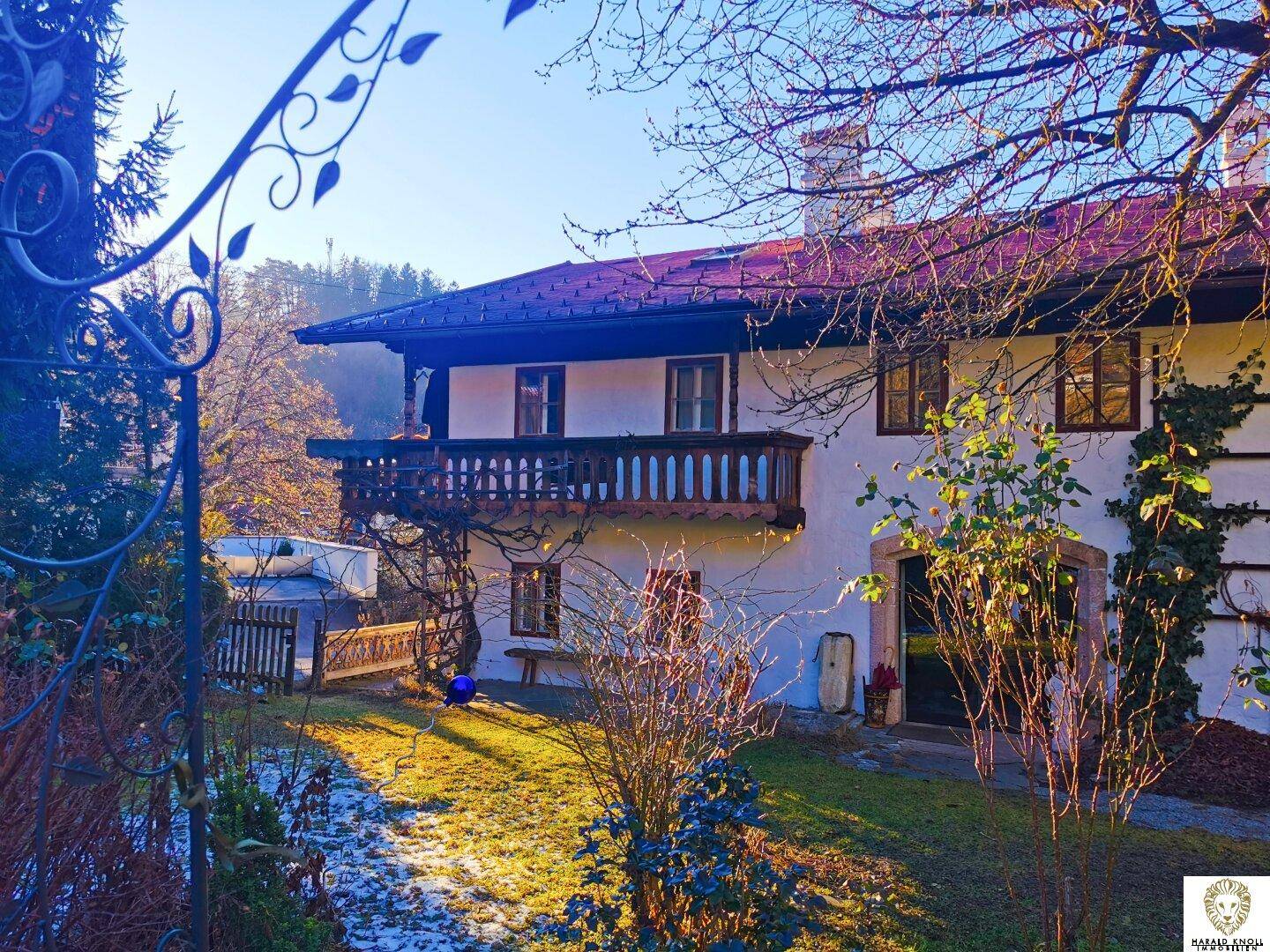 Alpenländisches Haus mit Holzbalkon und gepflegtem Garten im Sonnenlicht, umgeben von Bäumen.
