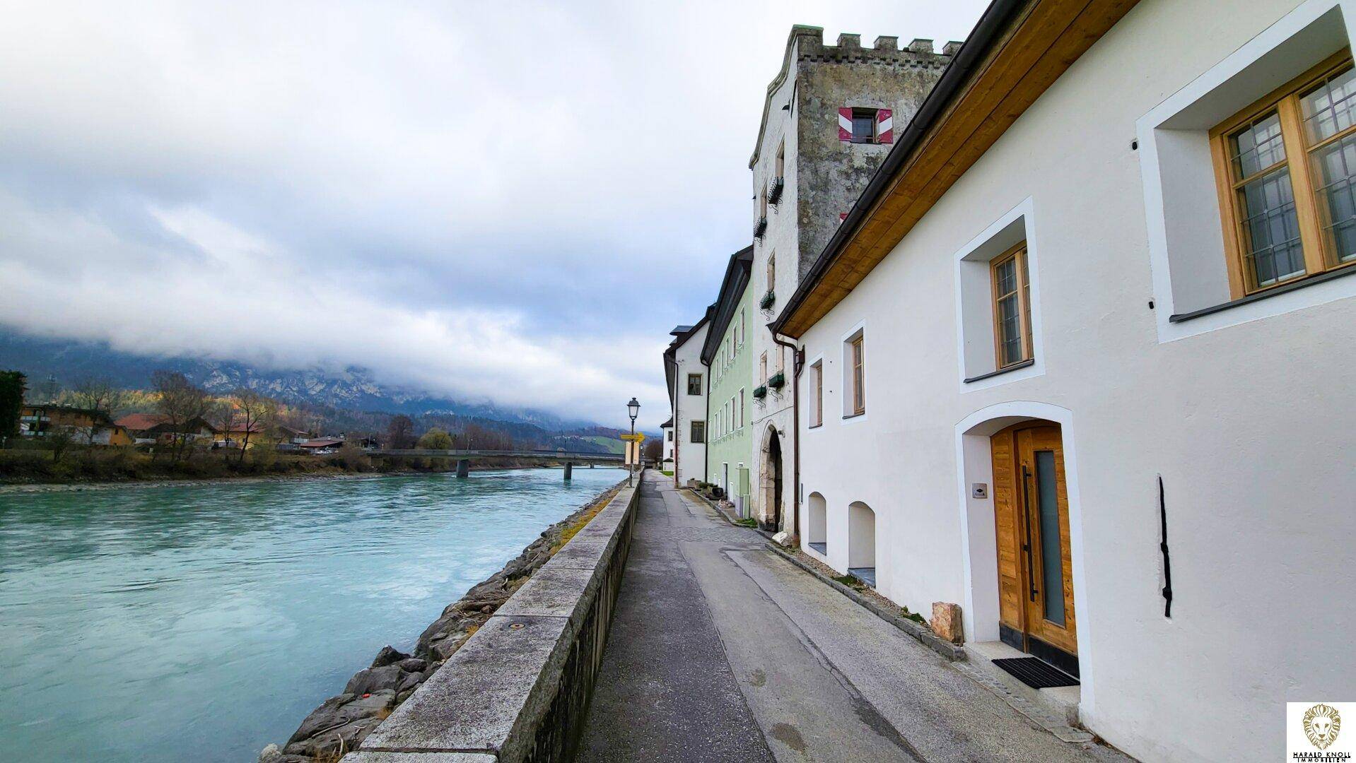 Uferpromenade mit historischen Gebäuden, einem Fluss und einer Brücke im Hintergrund, umgeben von Bergen.