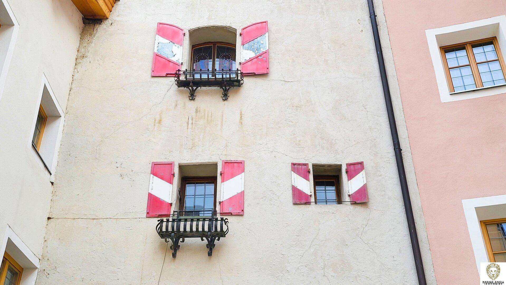 Fassadendetail mit Fenstern, die rot-weiß gestreifte Fensterläden und schmiedeeiserne Blumenkästen zeigen.