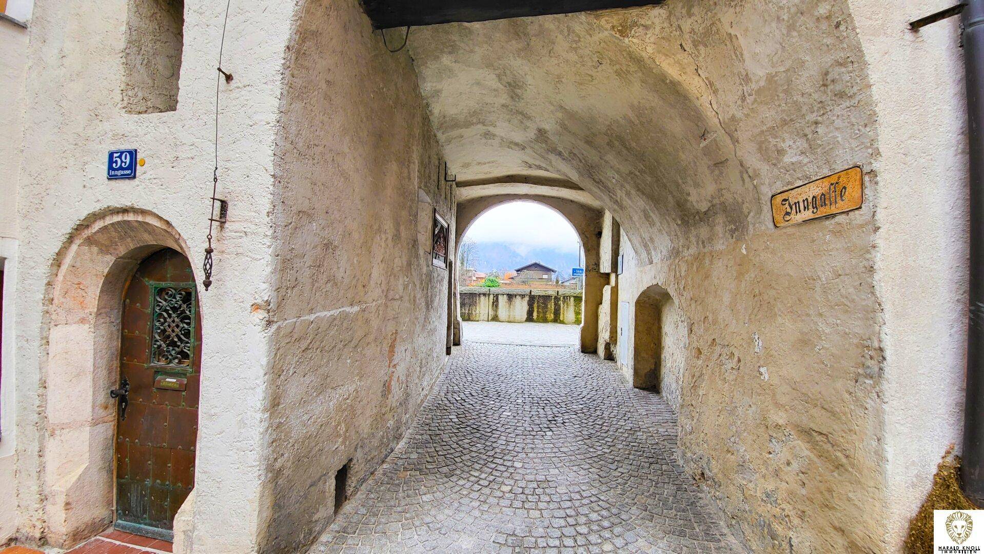 Historischer Torbogen mit gepflastertem Durchgang und einer alten Holztür, die zu einem offenen Platz führt.
