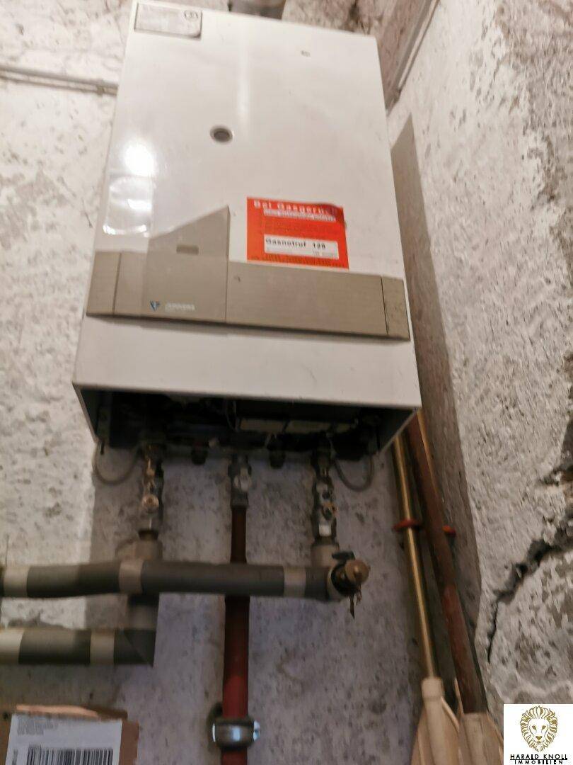 Wandmontierter Gasboiler oder Warmwasserbereiter in einem einfachen Technikraum mit rauen Wänden.