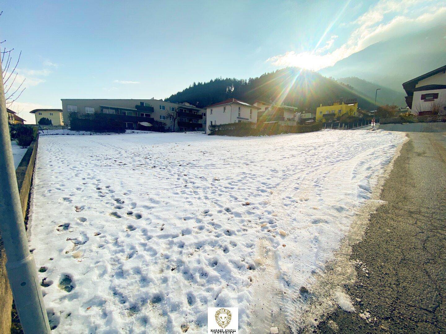 Sonniges, schneebedecktes Grundstück mit Bergblick und umliegender Bebauung unter blauem Himmel.