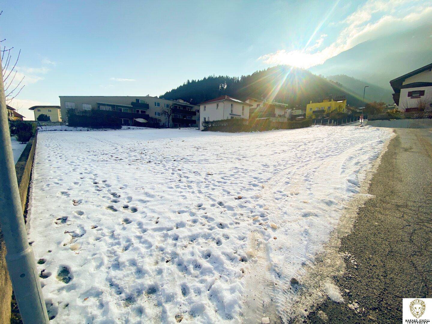 Sonnenbeschienenes, schneebedecktes Grundstück mit Bergen im Hintergrund und umliegender Bebauung.