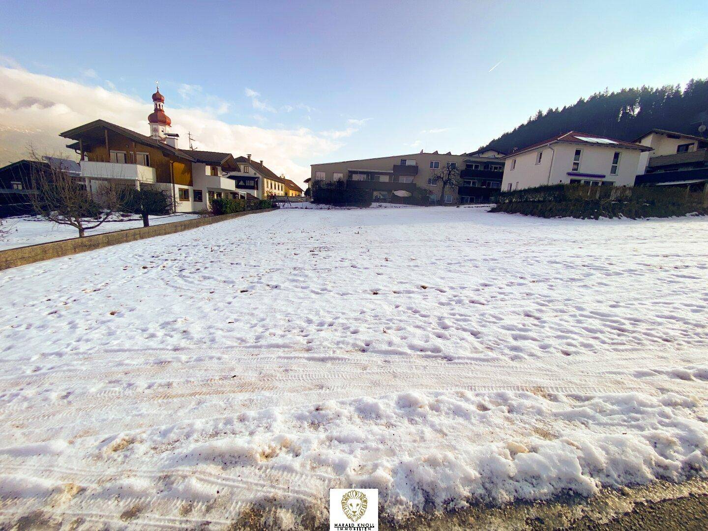 Breiter Blick auf ein schneebedecktes Grundstück, umgeben von Wohngebäuden und einem markanten Kirchturm.