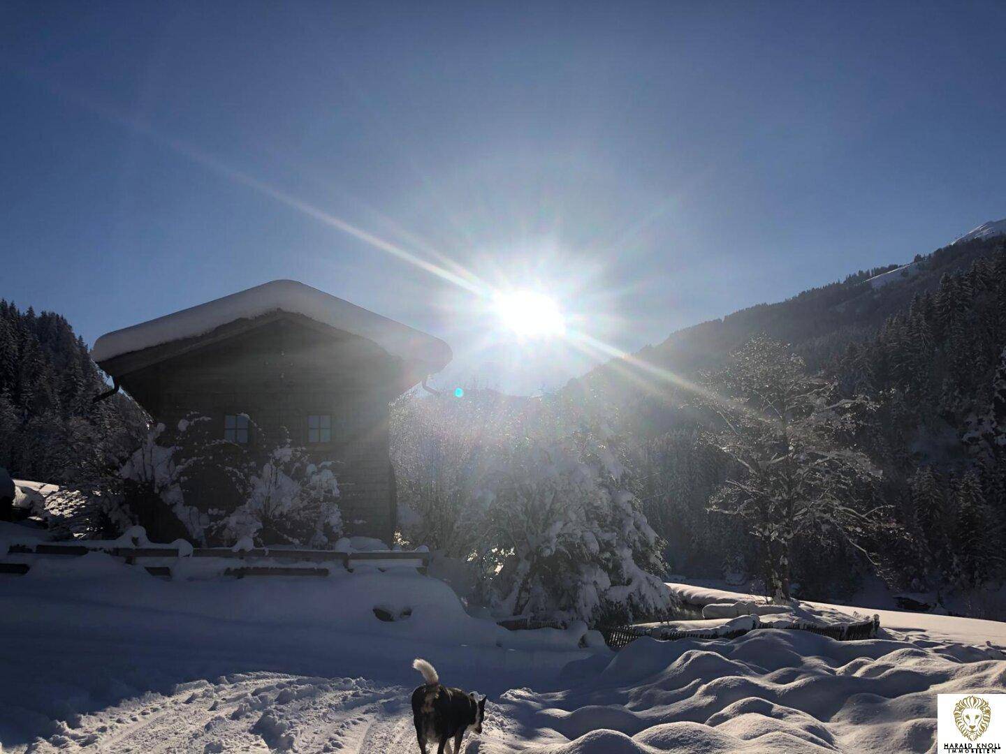 Winterlandschaft mit verschneitem Holzhaus und strahlendem Sonnenschein über den Bergen.