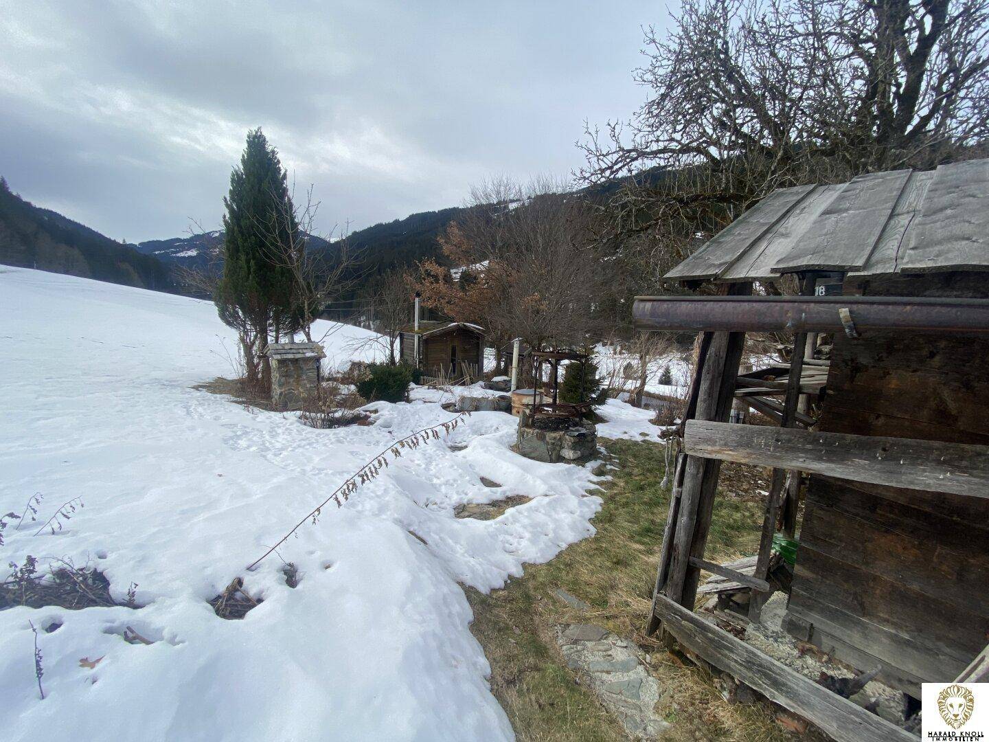 Verschneite Landschaft mit einem kleinen Holzschuppen und Blick auf die umliegenden Berge.