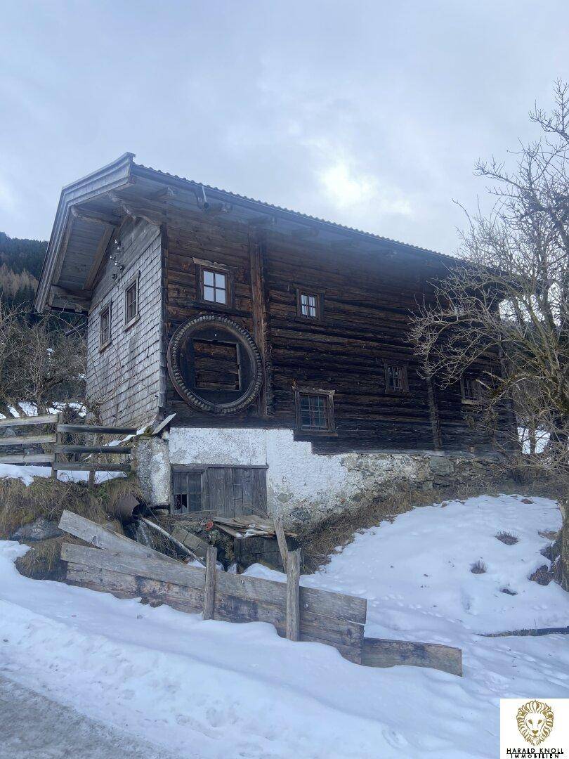 Rustikales Holzhaus mit altem Wasserrad und verschneitem Hang im Vordergrund.
