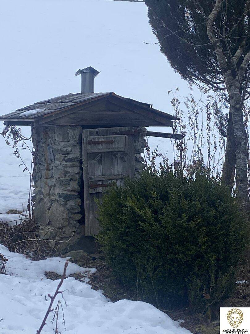 Einfaches Steinhäuschen mit Holztür und Schornstein, umgeben von Schnee und Büschen.