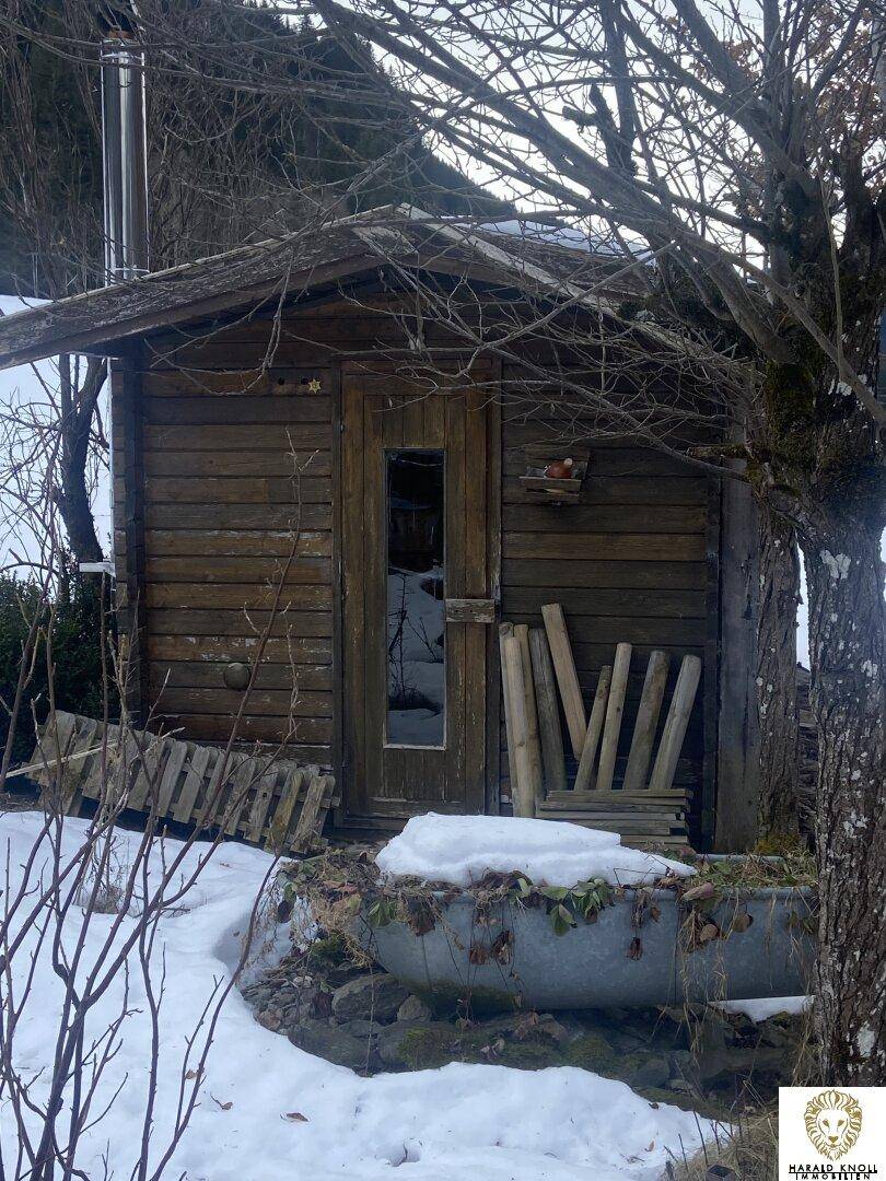 Kleiner Holzschuppen mit Tür und Fenster, umgeben von Schnee und kahlen Bäumen.
