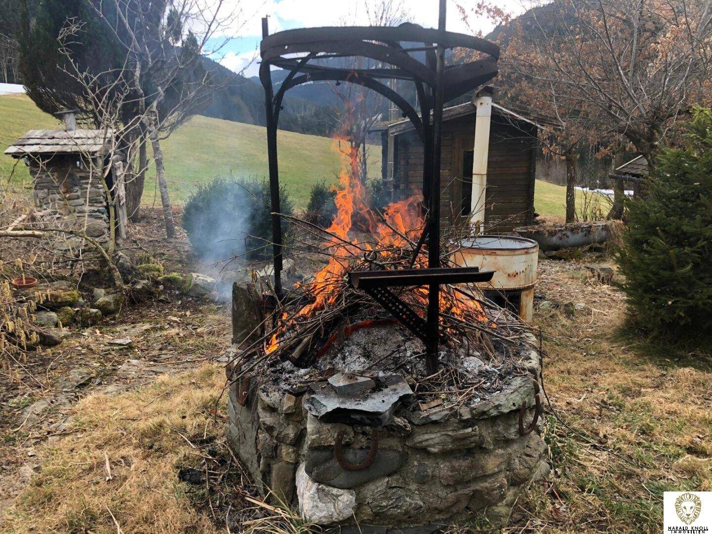 Gemütlicher Feuerplatz aus Stein mit brennendem Holz und rustikaler Metallkonstruktion.