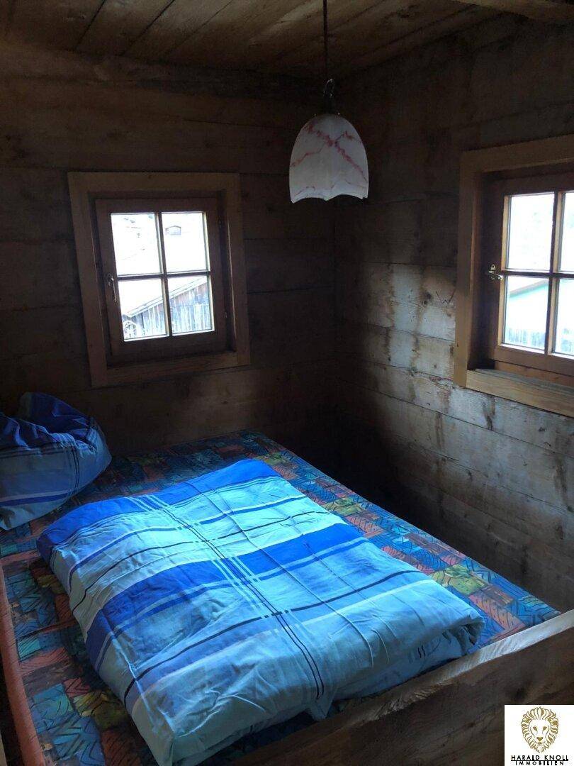 Gemütliches Schlafzimmer mit Holzwänden, Holzboden und einem Bett mit blauer Bettwäsche, beleuchtet durch zwei Fenster.