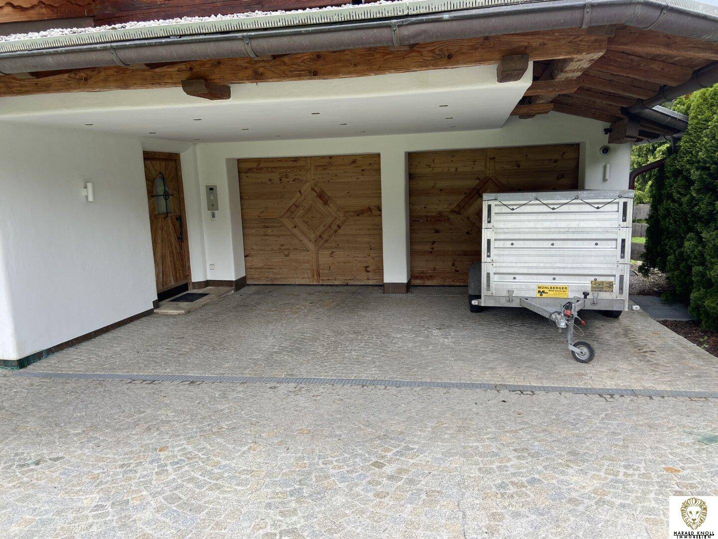 Überdachter Eingangsbereich mit zwei Holzgaragentoren und gepflasterter Zufahrt.