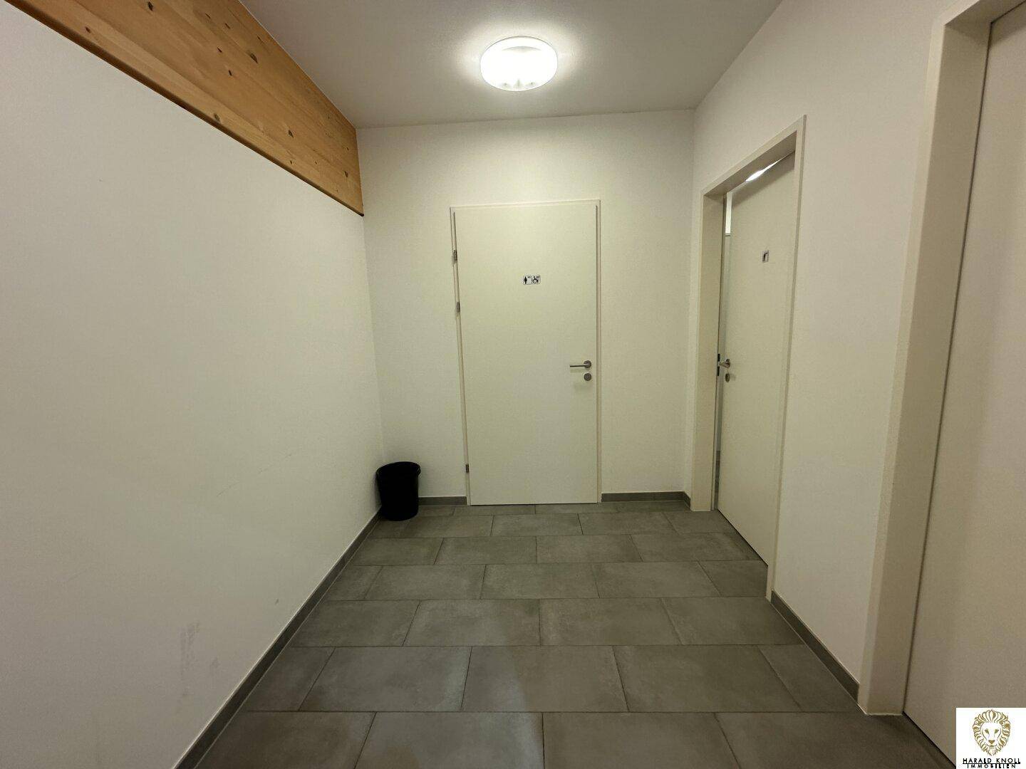 Eingangsbereich zu den Toiletten mit grauen Fliesen und Holzbalken an der Decke.