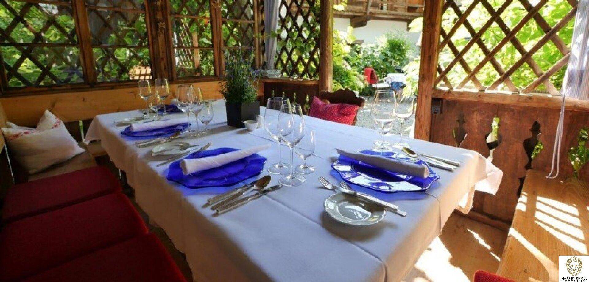 Überdachte Terrasse mit Holzelementen, gedecktem Tisch und blauen Tellern, umgeben von viel Grün.
