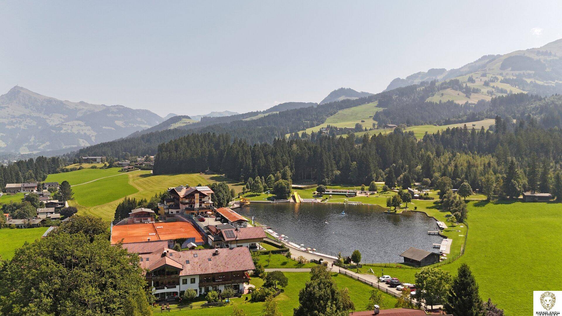 Panoramablick auf einen idyllischen See, umgeben von grünen Wiesen, Wäldern und majestätischen Bergen.