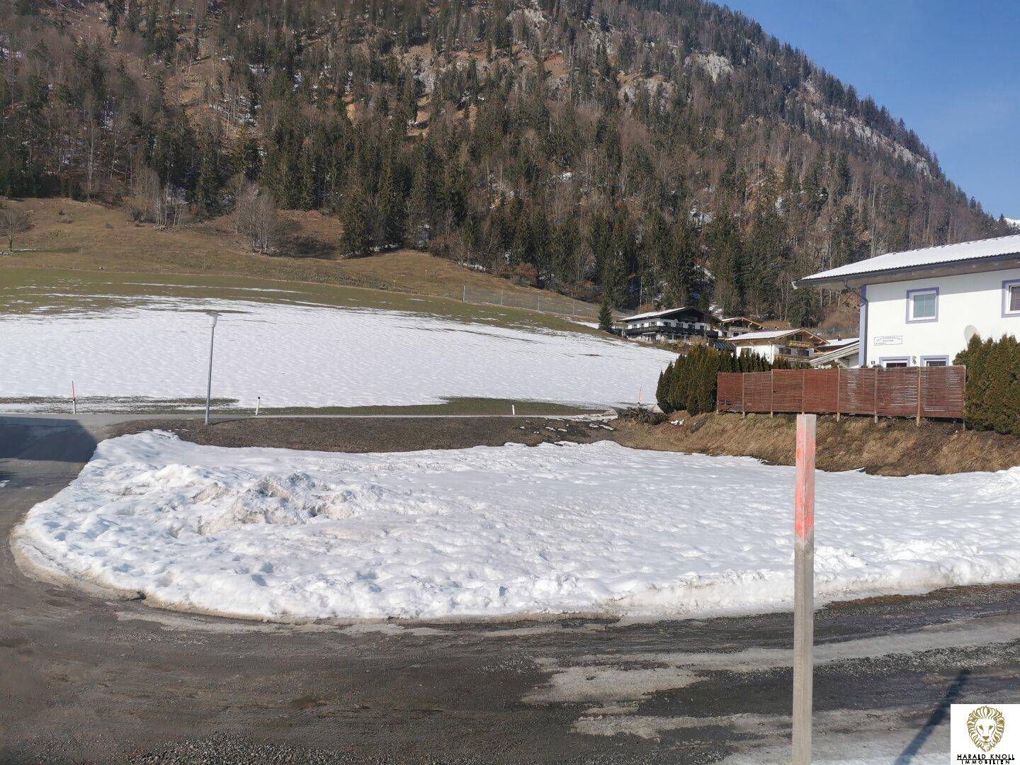Schneebedeckte Landschaft neben einer Straße mit bewaldeten Bergen im Hintergrund und vereinzelten Häusern.
