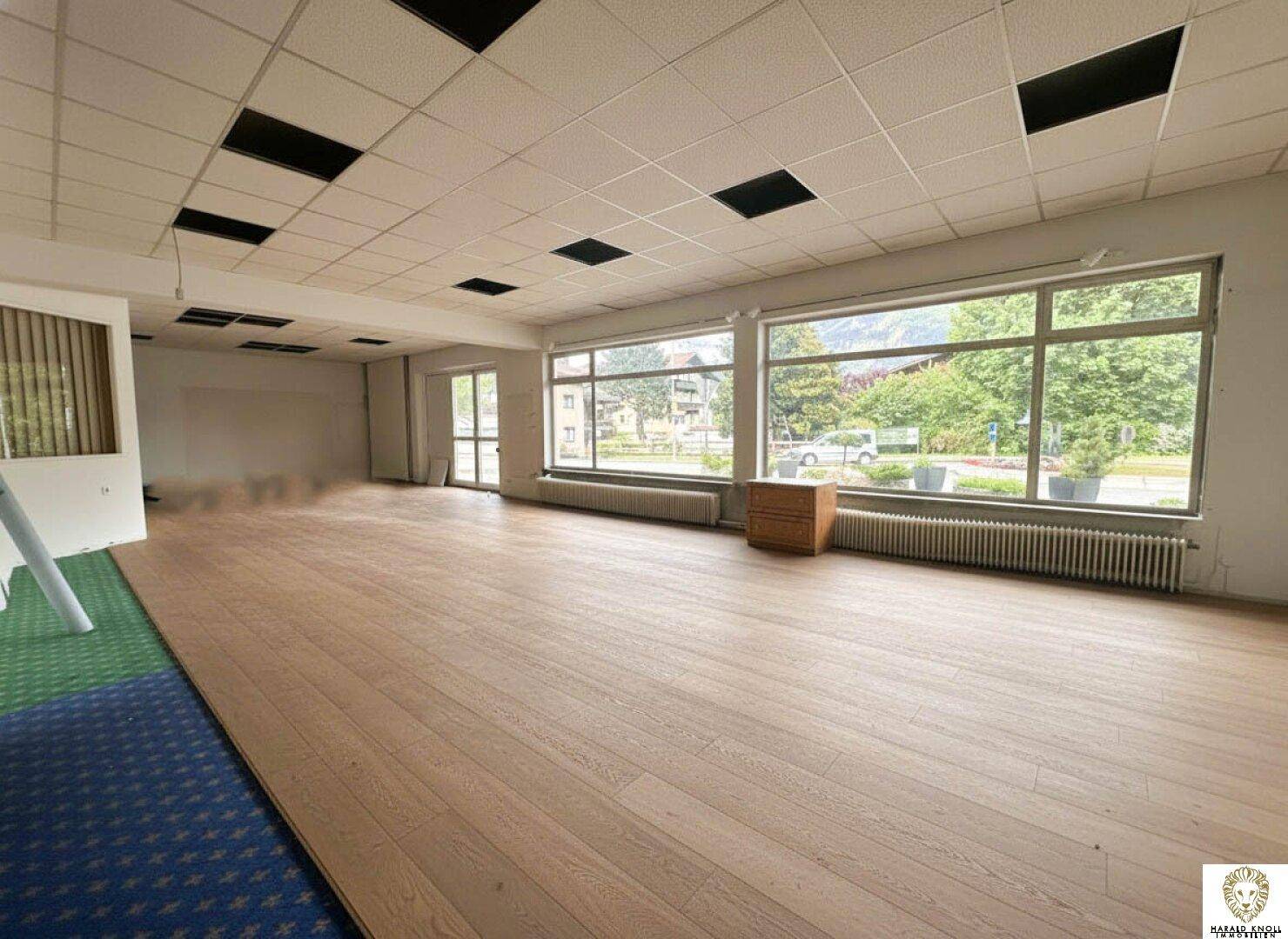 Geräumiger, leerer Geschäftsraum mit großen Fenstern, Holzboden und Teppichbereich.