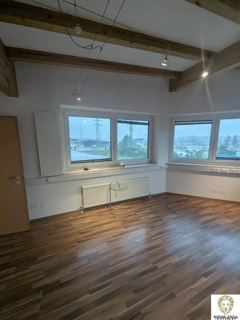 Geräumiges Büro mit sichtbaren Holzbalken, Laminatboden und Heizkörpern unter den Fenstern.