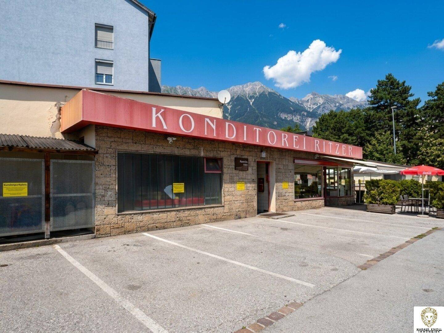 Fassade der Konditorei Ritze mit Parkplätzen davor und majestätischen Bergen im Hintergrund.