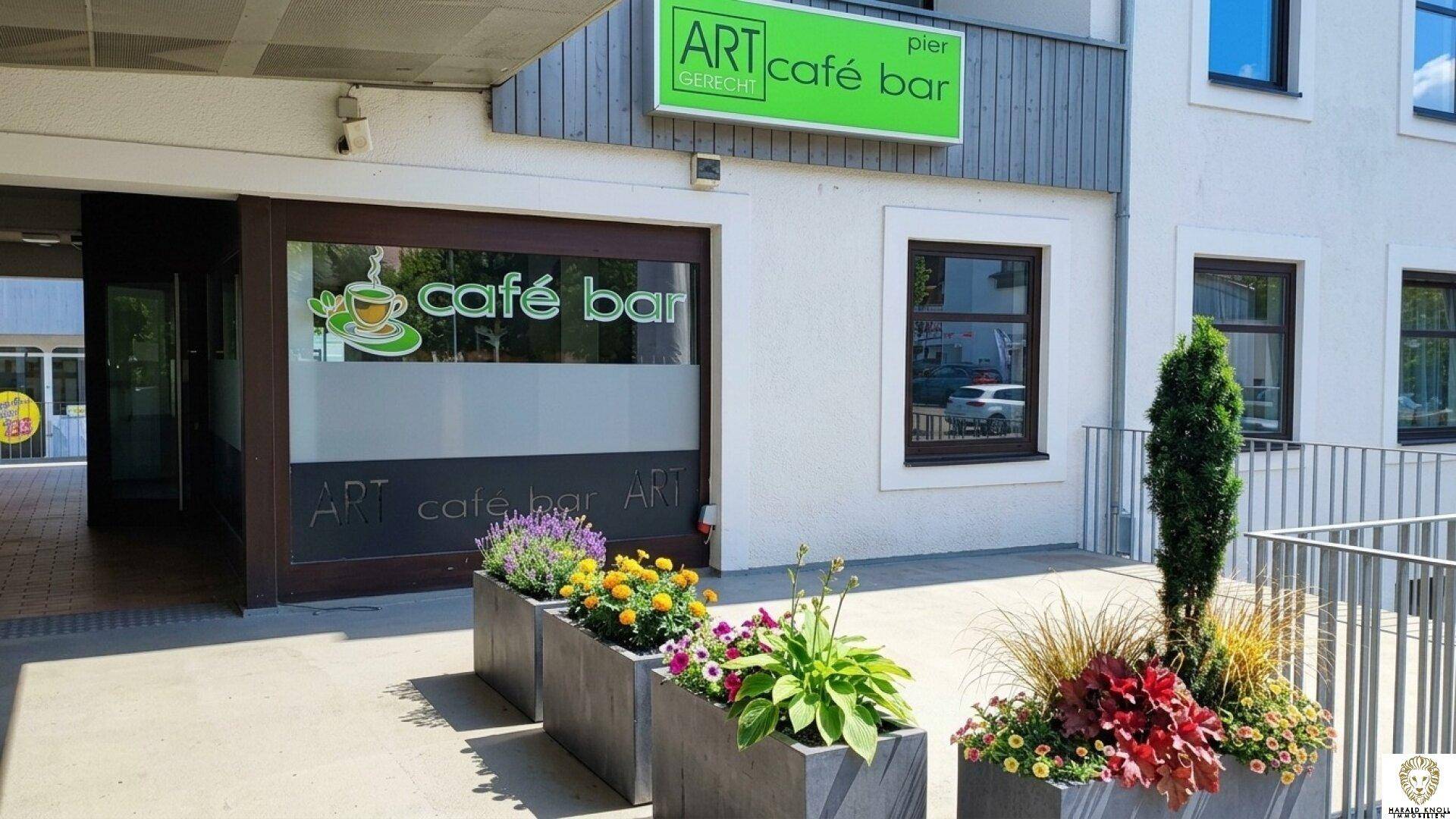 Eingangsbereich eines Cafés mit Blumenkübeln und einem Schild über der Tür.