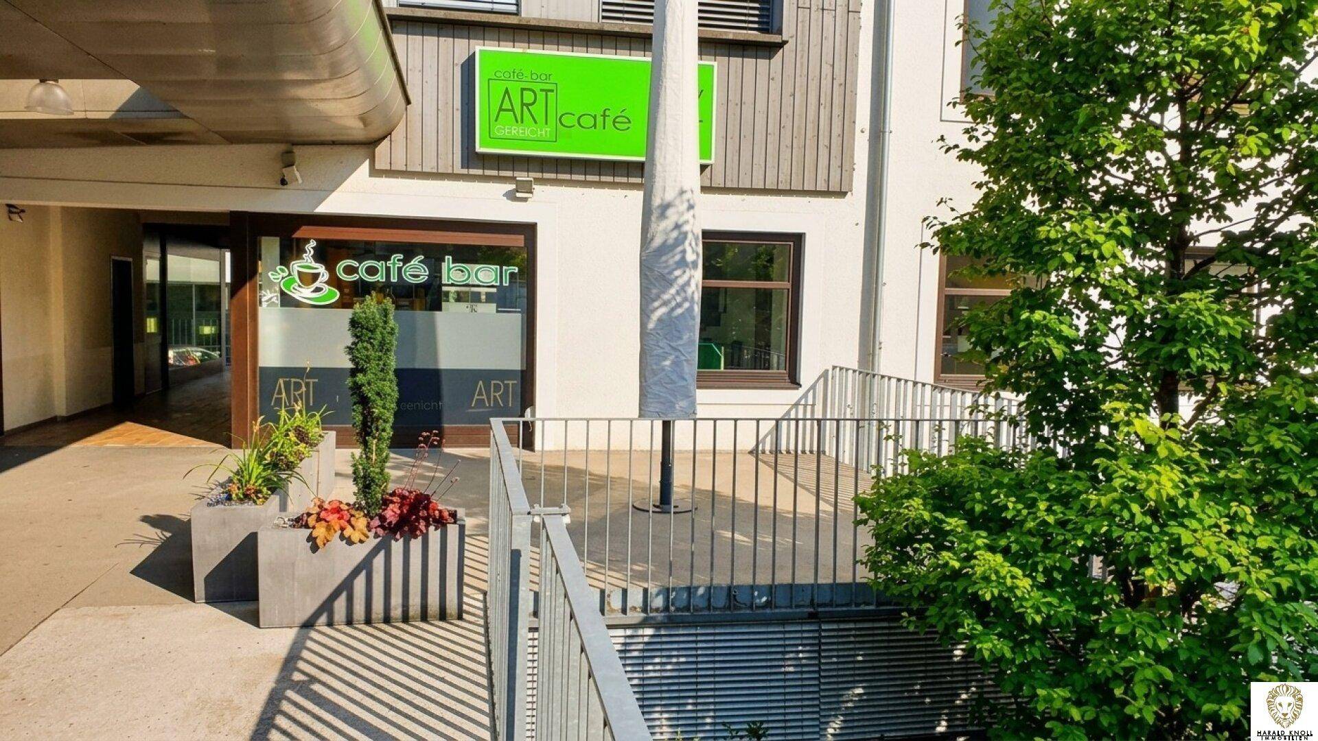 Außenansicht eines Cafés mit Treppengeländer, Pflanzen und einem großen Baum im Vordergrund.