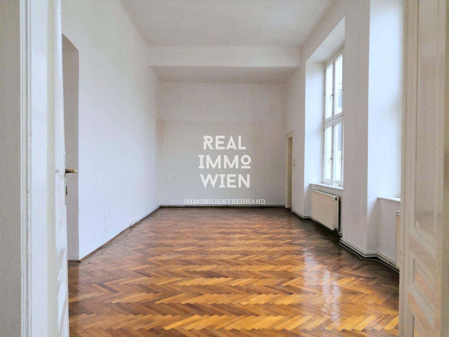 Leeres Zimmer mit glänzendem Fischgrätparkett, hohen Fenstern und Heizkörpern unter den Fensterbänken.
