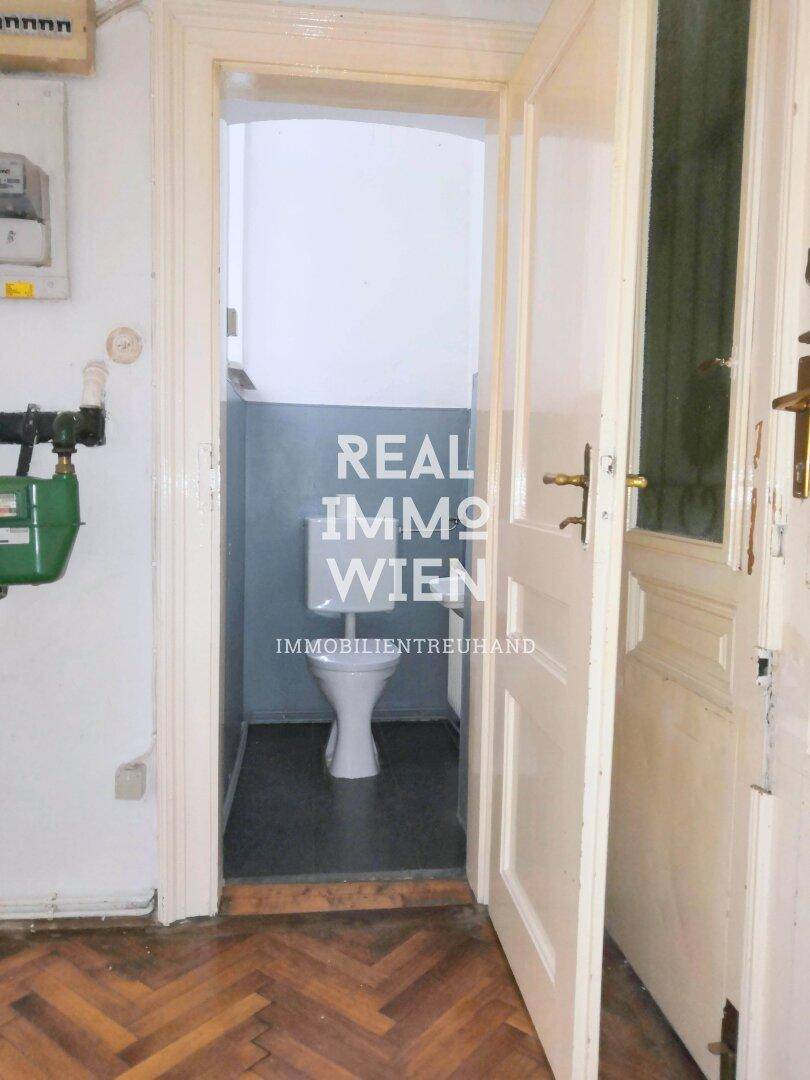 Kleiner Toilettenraum mit WC und dunklem Fliesenboden, zugänglich durch eine weiße Holztür.