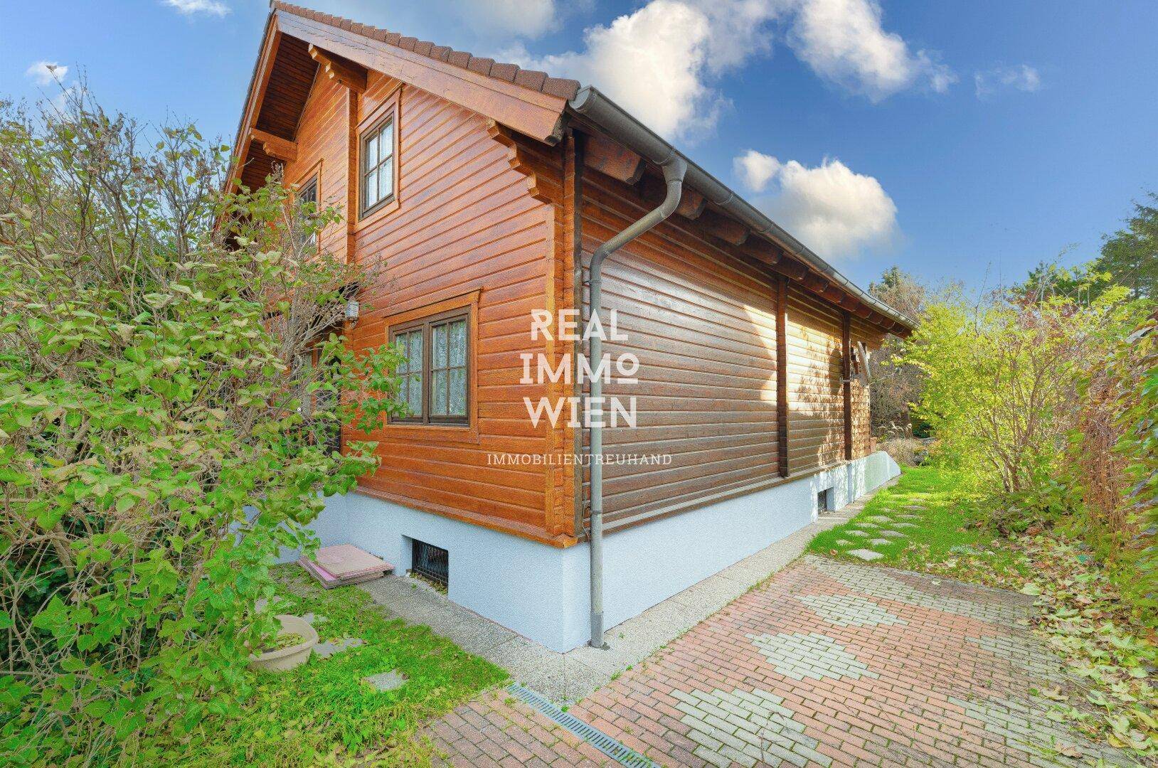 Seitenansicht des Holzhauses mit gepflasterter Auffahrt und grünen Büschen.