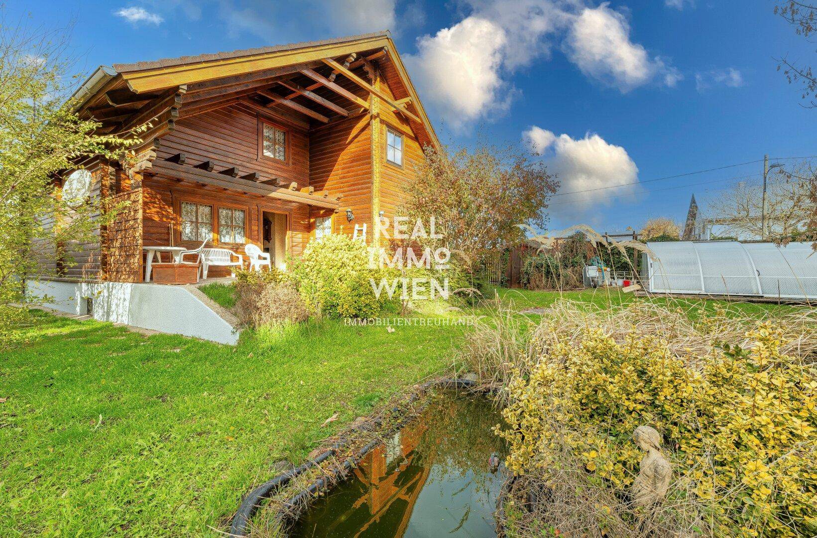 Idyllisches Holzhaus mit Teich im Vorgarten und üppiger Vegetation unter blauem Himmel.