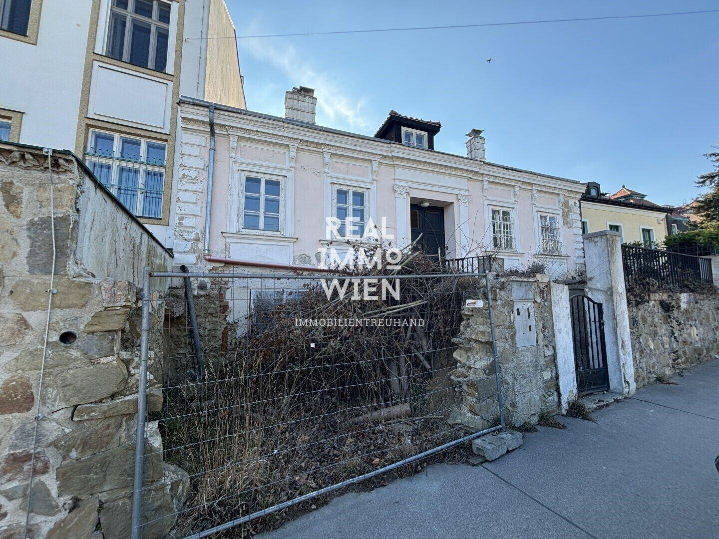 Historisches Wohnhaus mit verwildertem Vorgarten und alter Steinmauer an der Straße.