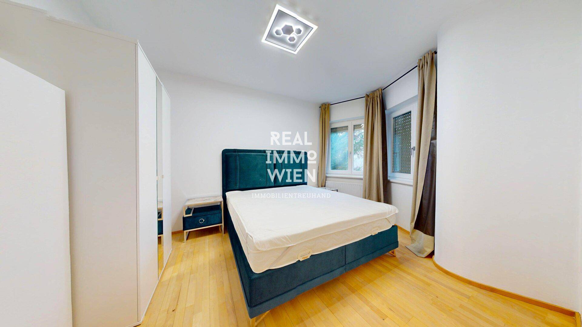 Schlafzimmer mit blauem Doppelbett, Holzboden und Fenster mit Blick ins Grüne.